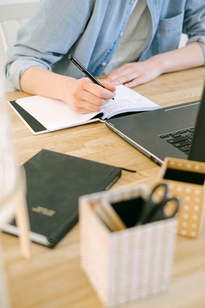 pexels-photo-4240497 Adult writing notes on a notebook while working on a laptop in a cozy home office environment.